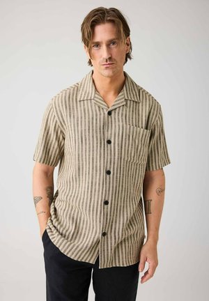Mann mit mittellangen Haaren, der ein beige-braun vertikal gestreiftes kurzärmeliges Hemd und schwarze Hosen trägt, steht vor einem einfarbigen Hintergrund.
