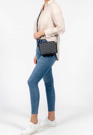 Zwarte schoudertas met een herhalend tekstpatroon, aangevuld met een ritsluiting en een verstelbare schouderband. Gedragen met een beige blouse en blauwe jeans.