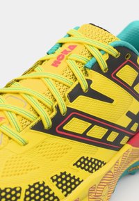 Zapatilla de running amarilla con parte superior de malla, acentos en negro y rojo, cordones turquesa, suela vibrante con patrón de lunares y un diseño texturizado.