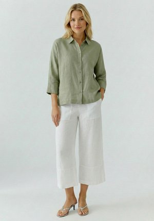 Femme portant une blouse vert clair boutonnée avec manches trois-quarts, un pantalon blanc large court, et des sandales argentées à talons, debout calmement.