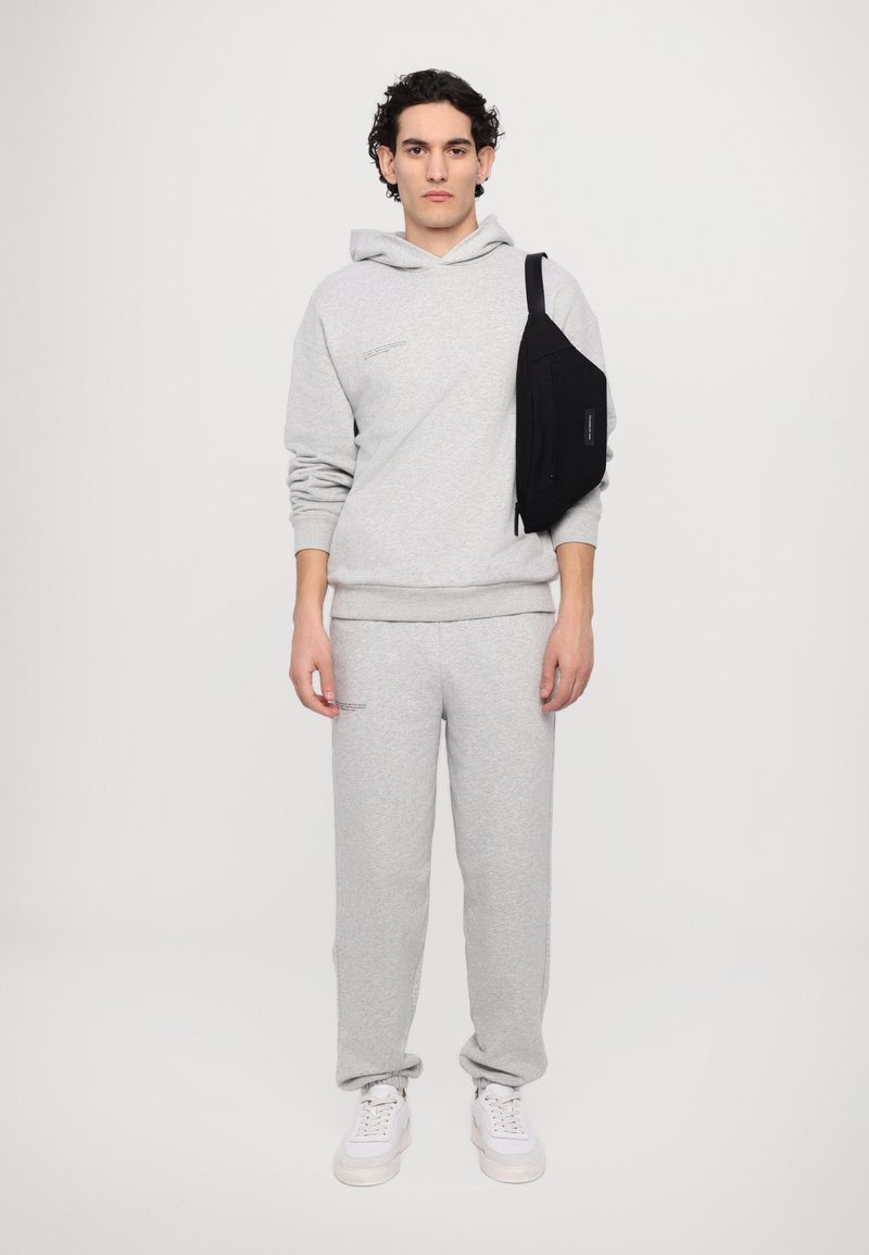 Pantaloni de trening gri și hanorac dintr-o țesătură moale, asortați cu adidași albi. O geantă neagră tip crossbody este purtată pe umărul stâng.