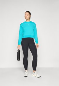Türkiser, gekürzter Pullover mit langen Ärmeln, schwarze Leggings und weiße Sneaker. Hält eine schwarze Wasserflasche. Einfaches, modernes Design.