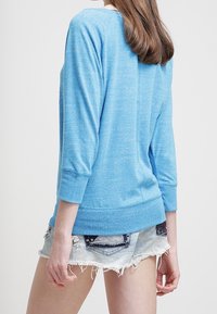 Ljusblå långärmad topp i mjukt material med avslappnad passform och uppvikta ärmar, matchad med slitna denimshorts.