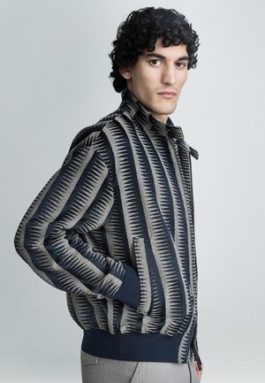 Blouson aviateur zippé avec un motif géométrique en bleu marine et beige. Confectionné en tissu texturé avec des poignets côtelés et un col montant.