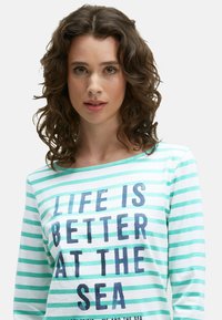 Junge Frau mit lockigem braunem Haar trägt ein gestreiftes Hemd in Weiß und Türkis mit der Aufschrift "Das Leben ist besser am Meer."