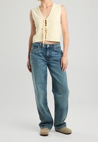 Krémový pletený top bez rukávů s hlubokým výstřihem do V a zavazováním, v kombinaci s modrými džíny s vysokým pasem a volným střihem.