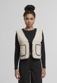 Krémová fleecová vesta s černými detaily, která má dvě velké přední kapsy a zavazování, nošena přes černý dlouhý rukáv.