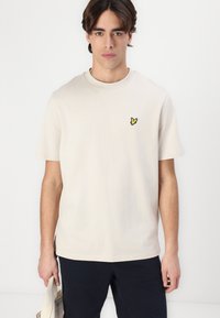 Beige bomulls t-skjorte med rund hals, korte ermer og en liten svart og gul brodert sommerfugl på brystet.