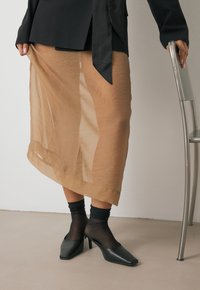 Läpinäkyvä vaaleanruskea hame pehmeällä tekstuurilla, yhdistettynä läpinäkyviin mustiin sukkiin ja kärjellisiin mustiin korkokenkiin. Minimalistinen muotoilu puhtain linjoin.