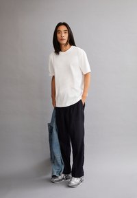 Witte T-shirt met korte mouwen, zwarte broek, houdt een spijkerjack vast. Grijze sneakers met reflecterende accenten. Neutrale achtergrond.