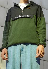 Zielony polarowy pulower z czarnymi panelami, krótkim zamkiem na ćwiartkę oraz dużym białym logo "ellesse". Zawiera pomarańczowy akcent logo i prążkowane mankiety.