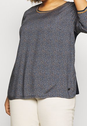 Vrouw draagt een donkerblauwe blouse met patroon en lange mouwen en beige broek, getoond van borst tot heupen tegen een witte achtergrond.