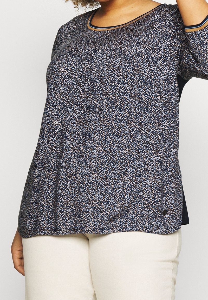 Femme portant un chemisier bleu foncé à manches longues avec motif, et un pantalon beige, montrée de la poitrine aux hanches sur un fond blanc.