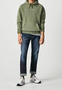 Sudadera con capucha verde oliva y bolsillo frontal; jeans azul oscuro con puños doblados; zapatillas blancas con acentos en rojo y azul marino.