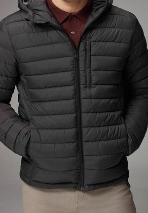 Mann trägt schwarze gesteppte Jacke mit Reißverschluss und Kapuze sowie ein bordeauxrotes Poloshirt, Hände in den Jackentaschen.