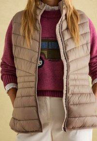 Beige Puffer-Weste mit horizontaler Steppung, Reißverschluss und hohem Kragen; getragen über einem rosa Strickpullover mit einem Autodesign.
