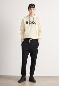 Jonge man in een crème hoodie met de tekst "BOSS", zwarte joggers met een trekkoord, zwarte pet en zwarte sneakers, staand tegen een effen achtergrond.