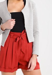 Veste gris clair texturée au design ajusté, associée à un short rouge taille haute plissé avec une ceinture nouée. Haut noir en-dessous.