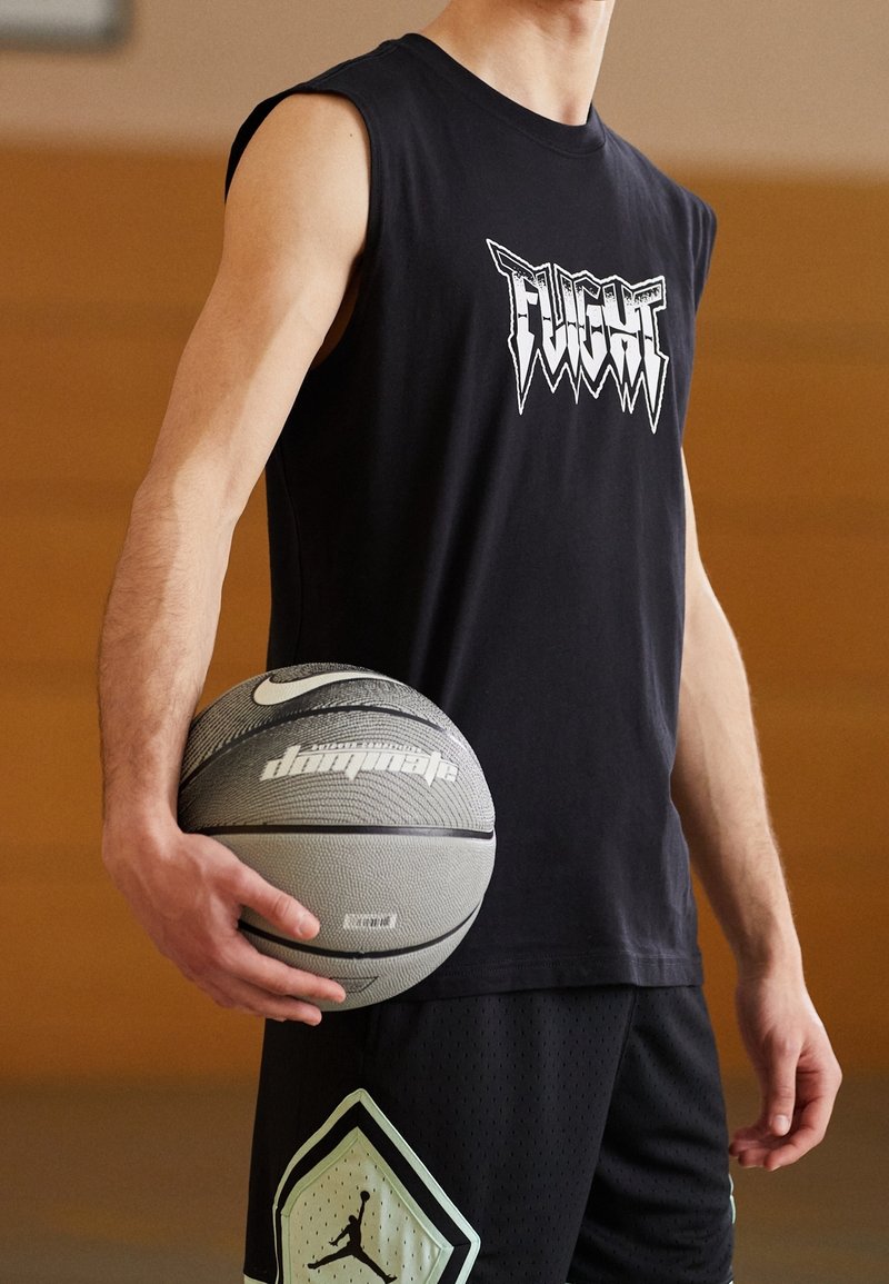 Personne en débardeur noir et short de basket tenant un ballon de basket Nike gris et noir sur un terrain intérieur.
