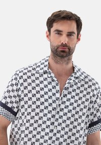 Camicetta a maniche corte di colore bianco, caratterizzata da un motivo geometrico nero e accenti solidi blu navy a contrasto sulle maniche. Dettaglio di chiusura a bottoni.