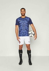 Puma spordipusa on tumepunase, mustriga disainiga; kaasas valged lühikesed püksid, mustad sokid ja must-valge jalgpall.