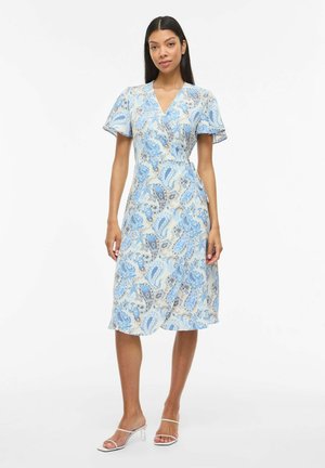 Frau trägt knielanges Wickelkleid mit blau-beigem Paisley-Muster und Schmetterlingsärmeln, steht in weißen Sandalen mit Absatz.