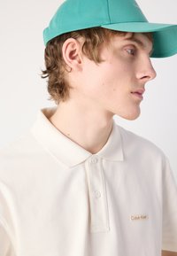 Bežs polo krekls ar klasisku apkakli, kurā ir mazs, brūns Calvin Klein logo. Pievienota ir tirkīza beisbola cepure.