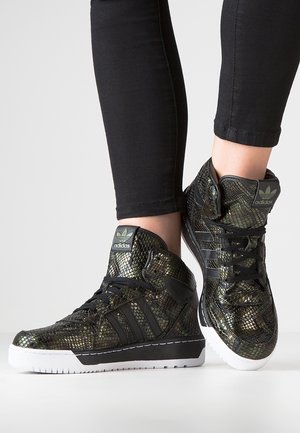 Czarne i metaliczne sneakersy Adidas na wysokiej podeszwie z wzorem skóry węża, noszone z czarnymi spodniami na jasnym, jednolitym tle.