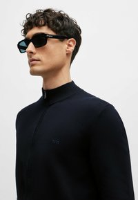 Maglione zip-up blu navy con colletto alto e logo "BOSS" discreto. Indossato con occhiali da sole neri angolari, che evidenziano un look elegante e moderno.