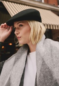 Schwarze Wollmütze mit runder Form, kombiniert mit einem weichen, grauen Schal in Deckenoptik. Der Hintergrund zeigt gedämpfte Töne mit gestreiften Markisen.