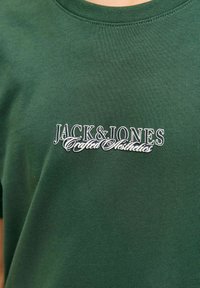 Groene katoenen T-shirt met een ronde hals en een wit logo met "JACK & JONES Crafted Aesthetics" op de voorkant gedrukt. Eenvoudige textuur.