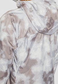 Lichte, geïsoleerde jas met een capuchon. Beschikt over een grijs en beige camouflagepatroon, gestructureerde stof en gathered elastische accenten.