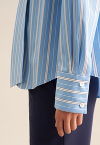 Camicia a righe azzurro chiaro con strisce verticali bianche, dotata di polsini con bottoni e una struttura liscia. Il tessuto è morbido e leggero.