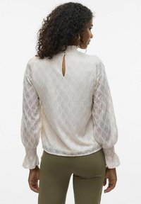 Witte blouse met textuurpatroon, hoge hals en sleutelgatopening aan de achterkant. Heeft pofmouwen met elastische manchetten.
