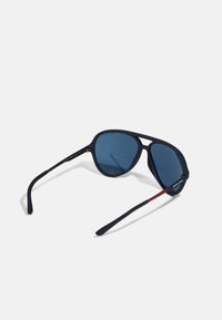 Ochelari de soare sport cu un cadru negru mat și lentile de un albastru închis. Caracteristicile includ brațe curbe ale templelor și un accent roșu subtil pe lateral.