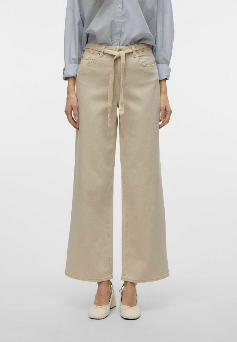 Beiga vida byxor i denim med framsluten snörning, sidofickor och en mjuk textur. Framträdande med sydda sömmar.