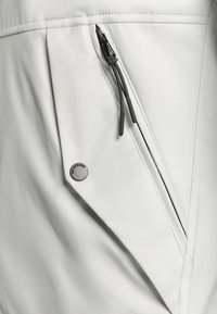 Witte, gladde buitenkleding met een ritszak met zwart trekband en een ronde metalen accent met de tekst "NO EXCESS."