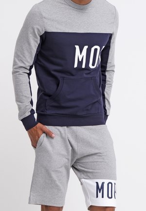 Homme portant un sweat gris et marine avec poche avant et un short gris assorti avec un texte marine sur un panneau blanc au-dessus du genou.
