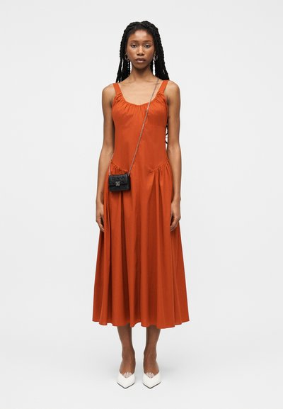 Γυναίκα με μακριά πλεξούδες, φορώντας πορτοκαλί μίντι φόρεμα χωρίς μανίκια, λευκές μυτερές γόβες, κρίκους σκουλαρίκια και μια μικρή μαύρη τσάντα χιαστί.