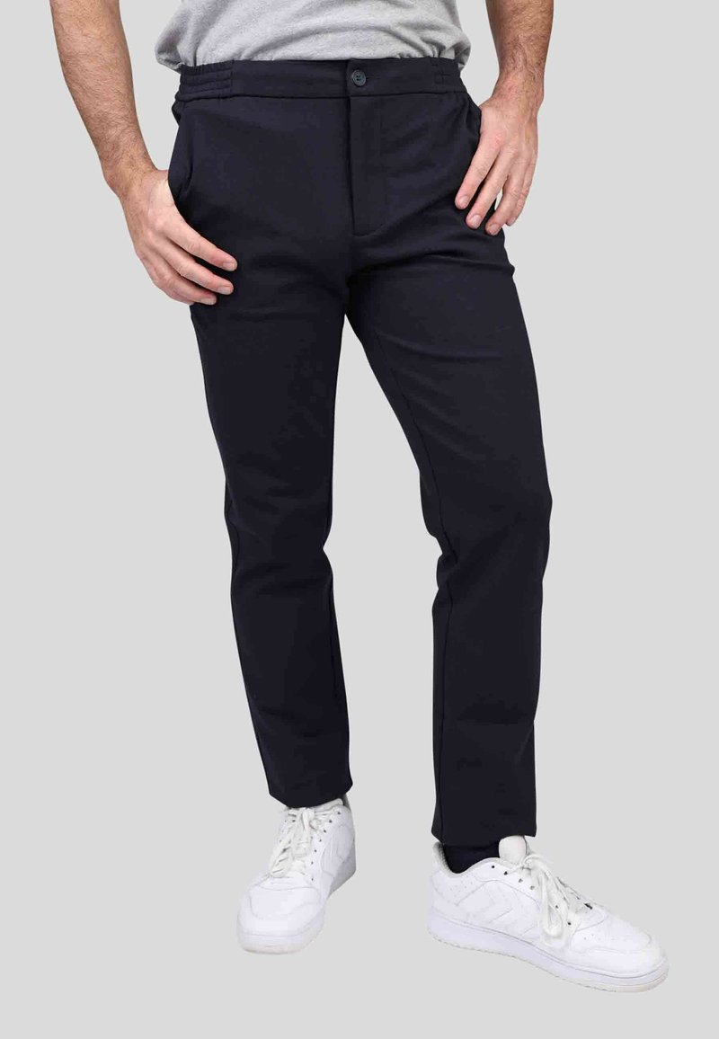 Pantalons ajustés navy avec une texture lisse, dotés de poches avant, de passants de ceinture et d'une fermeture à bouton, associés à des baskets blanches.
