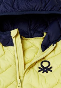 Chaqueta acolchada amarilla con un patrón acolchado, capucha azul marino y logo bordado azul marino en el pecho. Cuenta con un cierre frontal y tejido texturizado.