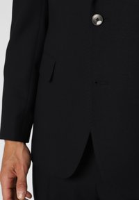 Dark blue blazer made of textured fabric, featuring a single button and a side pocket. Close-up view of the lapel and fabric detail.