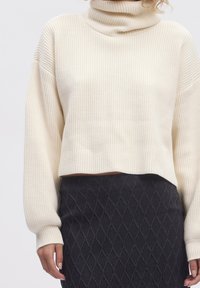 Pulover cremos cu guler înalt, cu curele lungi și largi. Asortat cu o fustă creion neagră texturată, cu un model în diamante.