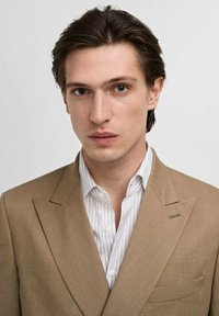 Giacca beige con rever a punta sopra una camicia bianca con sottili righe verticali. Tessuto morbido e vestibilità su misura con dettagli sui taschini.