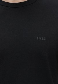 Personne portant un t-shirt noir uni à col rond avec le mot "BOSS" en petites lettres noires en relief sur la poitrine gauche.