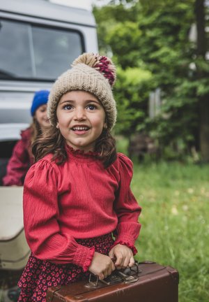 Rode textuur blouse met aandoenlijke mouwen, gecombineerd met een bloemenrok. Draagt een gebreide beige hoed met een roze pompon, en houdt een bruine koffer vast.