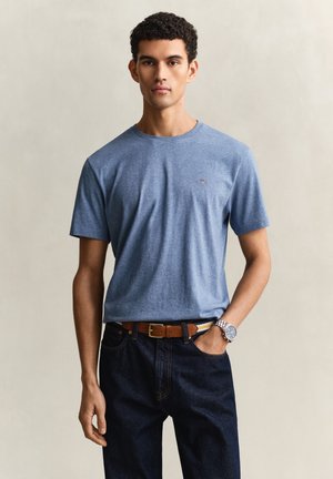 Junger Mann mit lockigem Haar trägt ein hellblaues T-Shirt mit kurzen Ärmeln, dunkle Jeans, einen braunen Ledergürtel und eine Armbanduhr und steht vor einem schlichten Hintergrund.