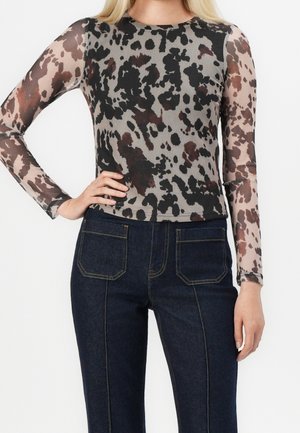 Kvinde iført en langærmet gennemsigtig top med sort og brune abstrakte pletter og mørkeblå denimjeans med forlommer.