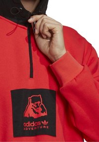 Pullover in pile rosso con cappuccio nero in contrasto. Presenta una tasca frontale con un grafico di un orso e il testo "adidas ADVENTURE" in grassetto. Chiusura a mezza zip.