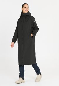 Zwarte, lange waterdichte jas met een kap, voorzien van een rits aan de voorkant, twee zijzakken en een relaxed fit. Gedragen over blauwe jeans en lichte laarzen.